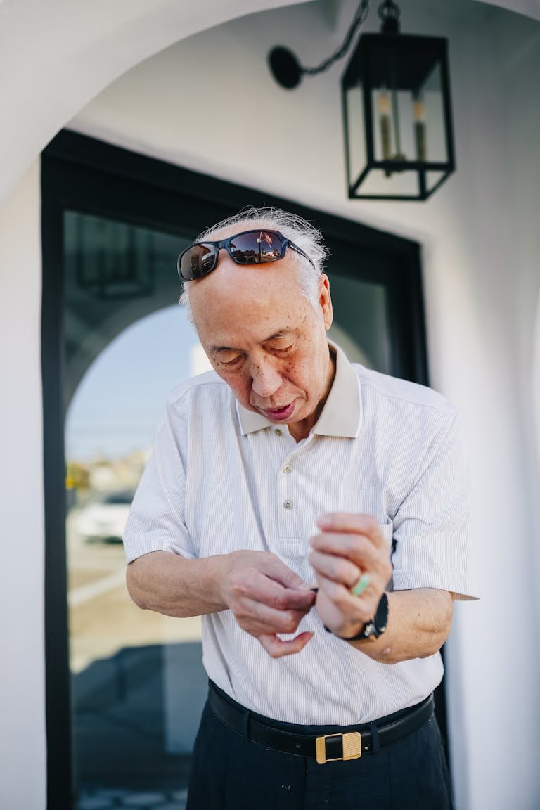 An elderly man wearing sunglasses adjusts his wristwatch outside a building.